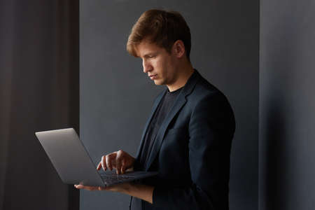 Profile portrait of a handsome young businessman in jacket, holding in hands a laptop, over black background.の写真素材