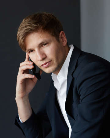 Young businessman in elegant suit sitting and looking at camera, holding his phone near his ear, over grey background.の写真素材