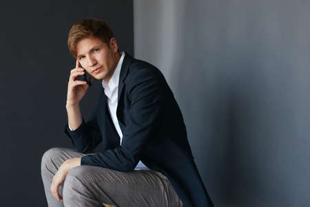 Handsome young businessman in suit sitting and looking at camera, holding his phone near his ear, over black background.の写真素材