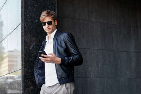 Confident young handsome business man wearing sunglasses, posing outside near building, read a message in his phone.の写真素材