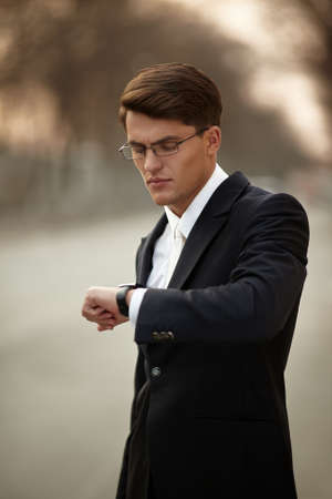 Portrait of an adult male model with glasses. Business man wearing black suit and white tie, looking at the his watch.の写真素材