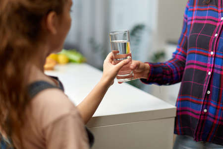 Frame image of a little girl holding a glass of water in the kitchen with the young mother.の写真素材