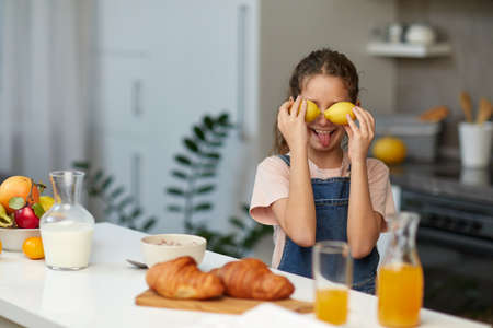 Little smiling cute girl holding of lemons near eyes and showing tongue over blurred kitchen background.の写真素材