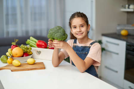Adorable little curly girl wearing jeans and t shirt, holding broccoli, seated at table in the kitchen.の写真素材