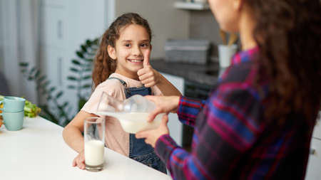 Closeup image of a mom pours her cute smiling daughter milk for breakfast, looking at camera shows thumb.の写真素材