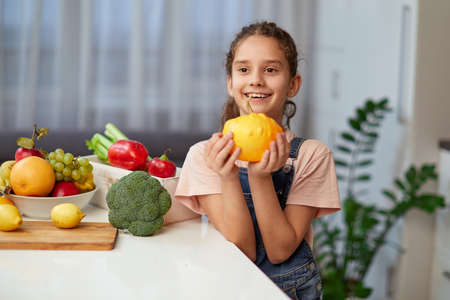 Front portrait of a cute little girl with curly hair, holding a yellow pumpkin, seated at table in the kitchen.の写真素材
