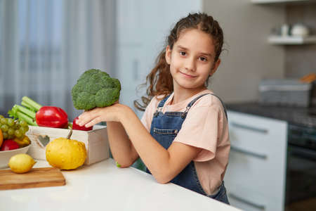 Front portrait of a little girl with curly hair, holding and looking at broccoli, seated at table in the kitchen.の写真素材