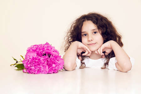 Pretty little girl with curly hair sitting at the table, looking at camera, have a pink bouquet of peonies.の写真素材