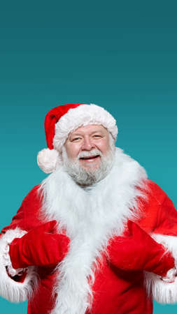 Closeup of a smiling Santa Claus in a red coat, gloves and a hat, with his hands raised high, isolated blue background.の写真素材