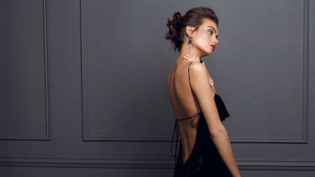 Rear view of a brunette woman in sexy black dress with bare back and beautiful earrings near grey dark wall.の写真素材