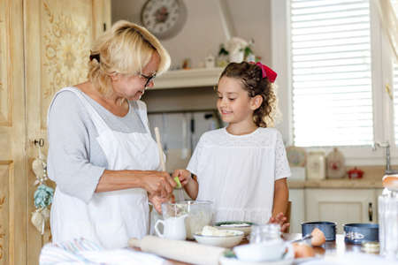 Grandmother and cute little curly granddaughter in the kitchen, they are preparing together homemade cookies.の写真素材