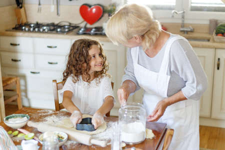 Adorable curly little girl with her grandmother cooking homemade cookies together at kitchen table.の写真素材