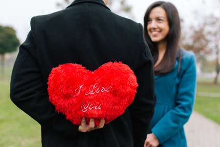 Concept of Valentines Day. Man standing and hiding red heart pillow behind his back. Girl looking at him and smiling.の写真素材