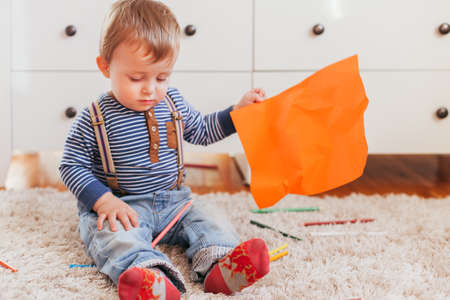 Cute little boy sitting on the floor and holding orange paper with color pencils around himの写真素材