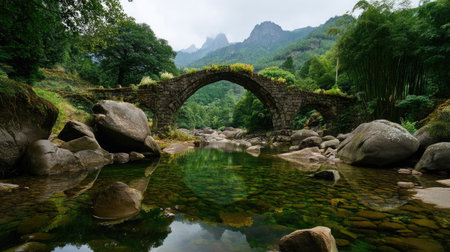 Capture the serene beauty of an ancient stone bridge arching over a calm river, surrounded by lush greenery and majestic mountains. Perfect for nature lovers.の素材