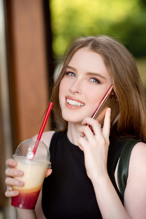 Young beautiful stylishly dressed girl is walking along the streets of the city, drinking juice, coffee, talking on mobile phoneの写真素材