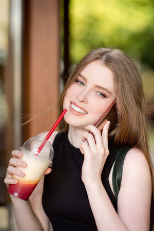 Young beautiful stylishly dressed girl is walking along the streets of the city, drinking juice, coffee, talking on mobile phoneの写真素材