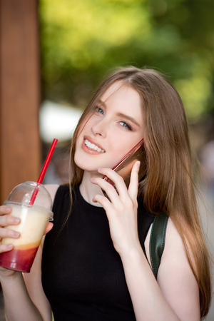 Young beautiful stylishly dressed girl is walking along the streets of the city, drinking juice, coffee, talking on mobile phoneの写真素材