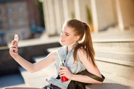 A beautiful young stylishly dressed girl walks in the city, drinks juice or a cocktail, eats sweets, makes selfie on a mobile phoneの写真素材