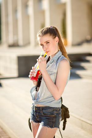 A beautiful young stylishly dressed girl walks in the city, drinks juice or a cocktailの写真素材