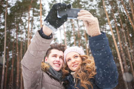 happy couple taking picture with smartphone selfie stick on over winter backgroundの写真素材