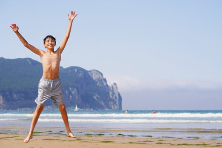 skinny teenage boy on the beach on vacation jumping happily in the sand with sea and mountain in the backgroundの写真素材
