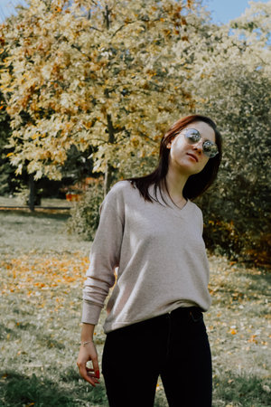 Young brunette girl in a beige sweater and black jeans posing in the autumn park.の写真素材