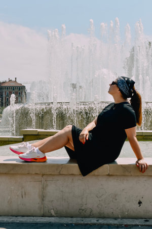 Brunette girl in black dress and sunglasses sitting near the fountainの写真素材