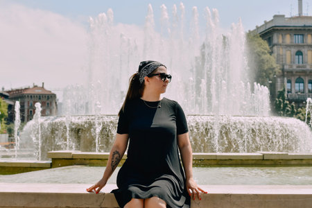 Young beautiful girl in a black dress and sunglasses sitting near a fountainの写真素材