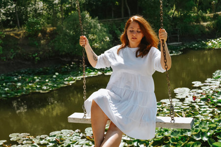 Redhead girl in white dress sitting on swing in the park.の写真素材