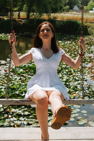 Young beautiful brunette woman in white dress on a swing in the parkの写真素材