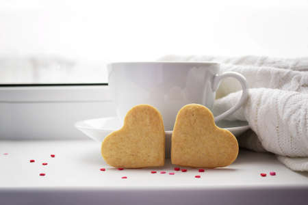 Two biscuits in the shape of a heart on a background with a cup of hot coffee and a blanket on the window.の写真素材