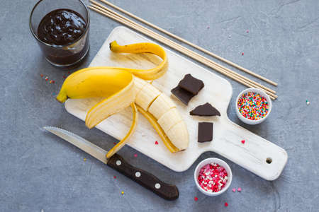 Cooking Banana Pops - bananas, dark chocolate, sweet sprinkling and cutting board, gray concrete background, top viewの写真素材