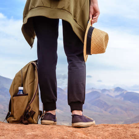 Girl tourist stands on hill. Legs of traveler, hat and backpack with bottle of water close-upの写真素材