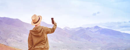 Girl tourist tourist in hat makes panorama of blue mountains on smart phone. Toning sunrise.の写真素材