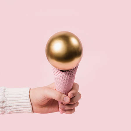 hand in white sweater holds pink ice cream cone with golden ball. Minimal Christmas concept.