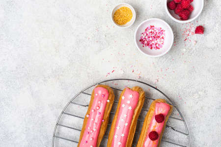 Delicious eclairs with pink glaze, raspberry and sugar sprinkles on gray background, copy space.