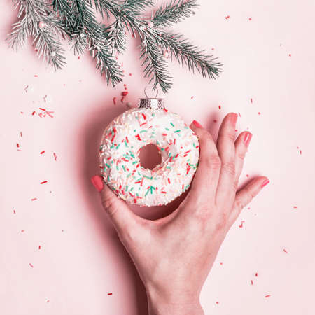 Female hand hangs Christmas bauble made of glazed donut on pink background. Christmas creative concept.の写真素材