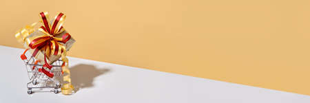 Shopping cart with gift box on beige gray background. Gifts wrapped in kraft paper with ribbon and bow. Holiday Shopping conceptの写真素材