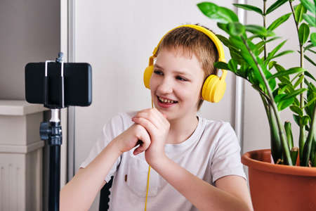 Boy blogger in yellow headphones talking with followers. Teenager live streaming looking to smartphone screen and gestures at homeの写真素材