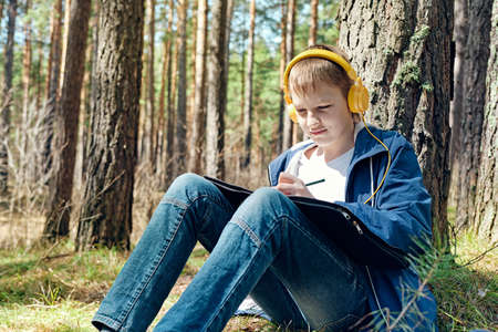 Boy sits in pine forest and plein air painting and listens to music.