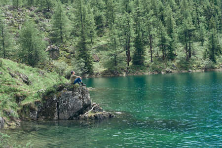 Alone man sits on a stone by the lake and looks at the beauty of nature. Calm and unity with nature