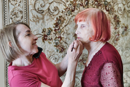 Elderly caucasian woman getting makeup applied by mature daughter in roomの写真素材
