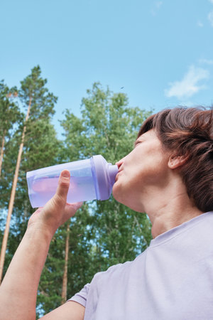 Mature woman drinks water from bottle after workout against background of forest and sky