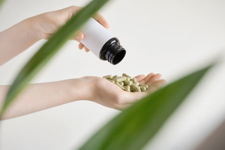 Female hands receiving herbal supplement capsules from bottle next to green plantの写真素材