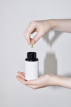 Hand holding a white blank label medicine bottle and adding green capsule
