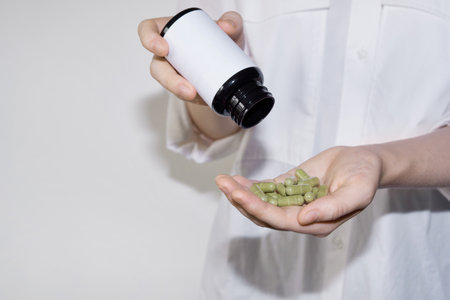 Hand holding green capsules while pouring from white supplement bottleの写真素材