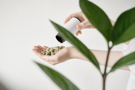 Female hands receiving green herbal supplement capsules from bottle next to green plantの写真素材