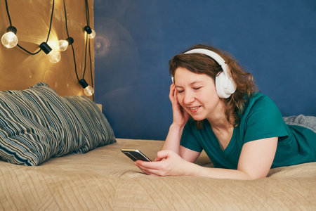 Woman lying on bed wearing headphones, smiling and using mobile phone