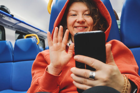Woman in hoodie traveling on train, waving and smiling during video call
